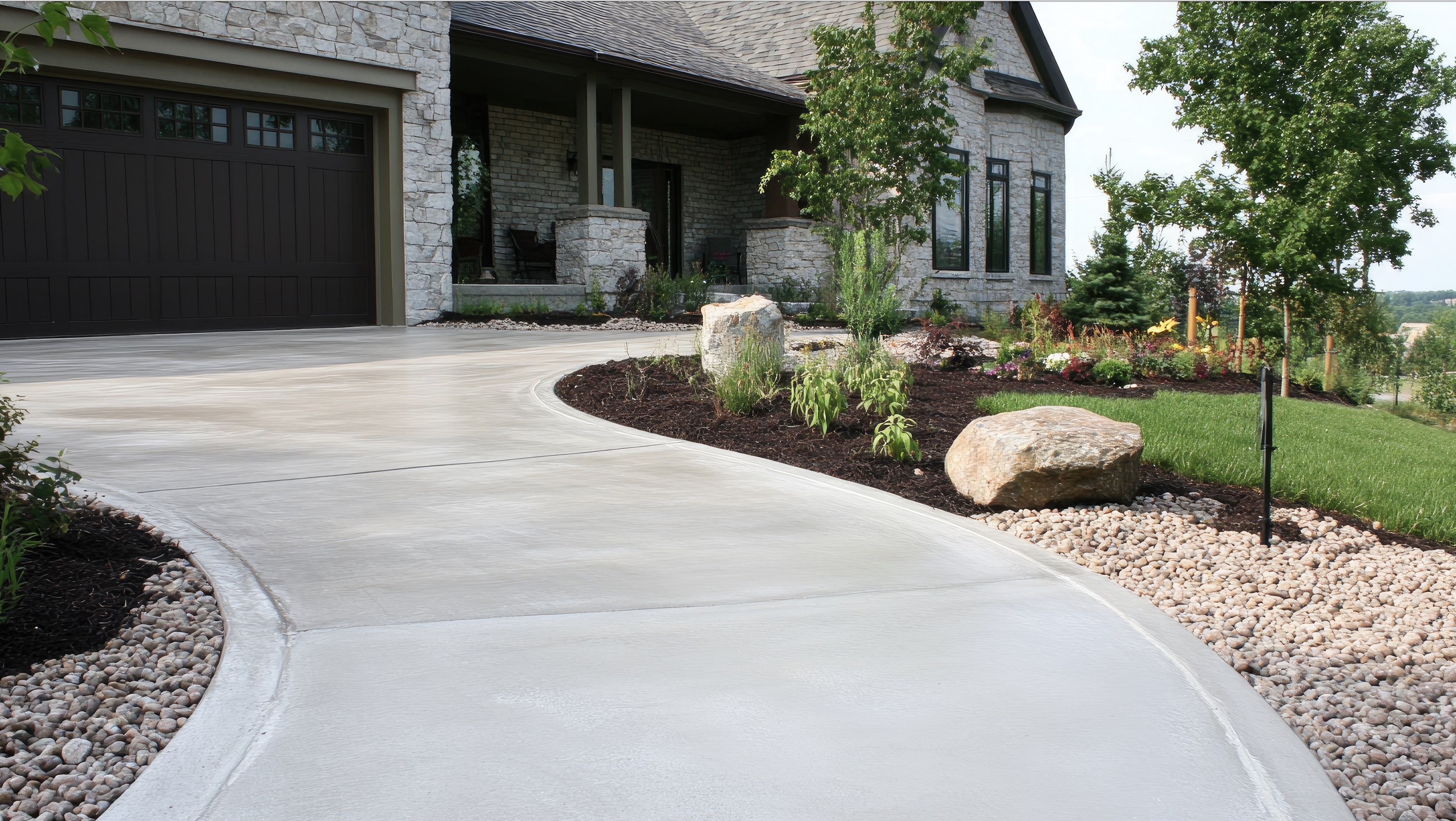 Finished concrete driveway curving up to a stone home, edged with rock and landscaping.