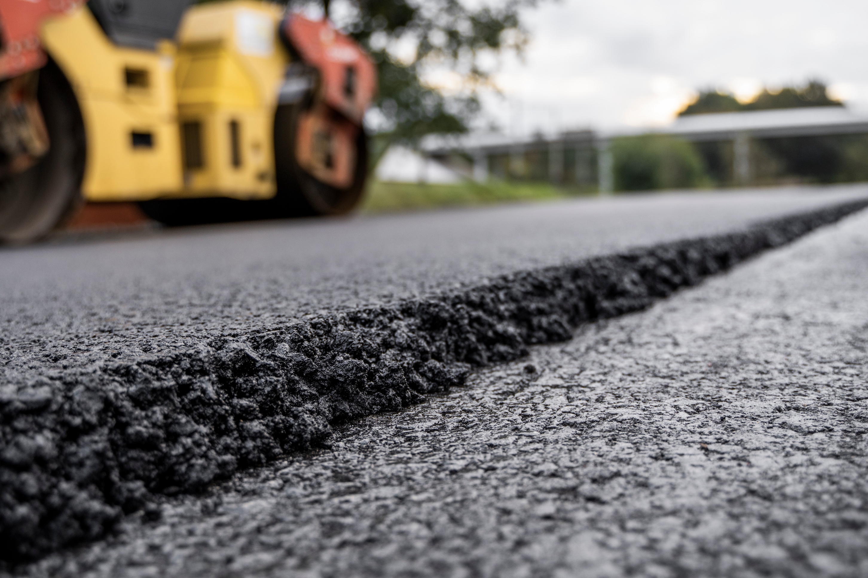 Close-up of fresh asphalt being laid and compacted by a roller, with the edge of the new road in sharp focus.